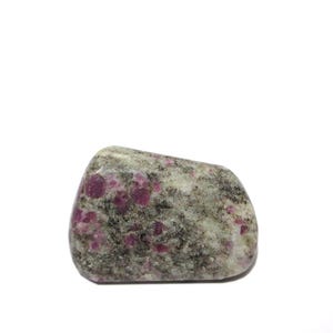 Puede incluir: Una piedra rodada gris con inclusiones rosas. La piedra tiene un aspecto moteado, con pequeños puntos rosas oscuros y redondos en toda su superficie. La forma es redondeada y lisa, lo que sugiere que ha sido pulida. El fondo es blanco.