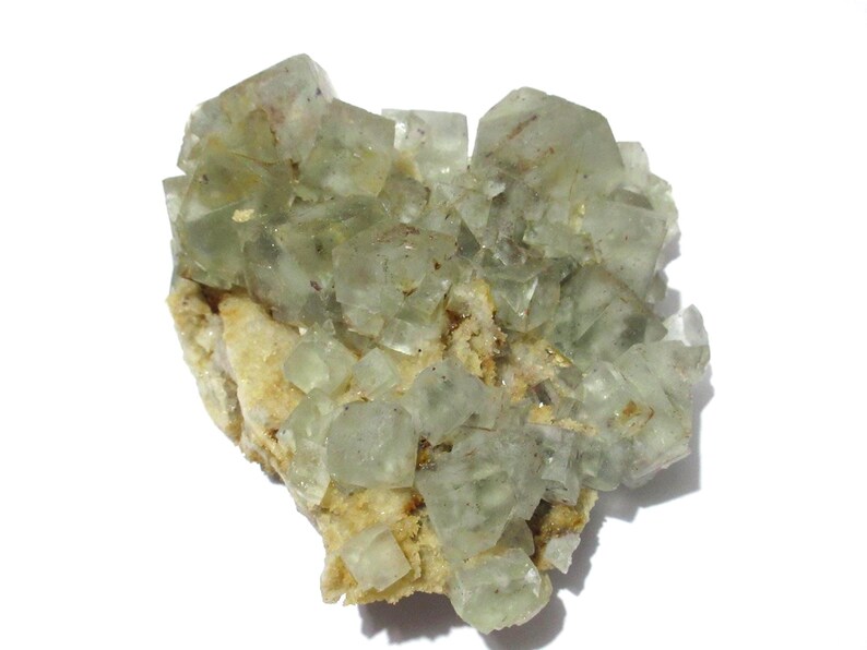 May include: A cluster of pale green fluorite crystals. The crystals are cubic in shape and sit atop a beige matrix. The fluorite has a translucent quality, and the overall appearance is natural and earthy. This mineral specimen is ideal for collectors.