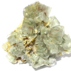May include: A cluster of translucent, pale green fluorite crystals. The crystals have a cubic structure and are arranged in a layered formation. The base is a light tan color, with some reddish-brown inclusions.