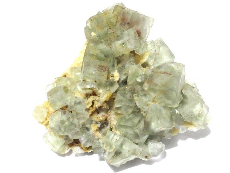 May include: A cluster of translucent, pale green fluorite crystals. The crystals have a cubic structure and are arranged in a layered formation. The base is a light tan color, with some reddish-brown inclusions.