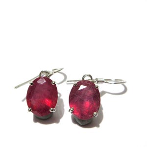 May include: A pair of ruby earrings. Each earring features a faceted, oval-shaped, deep red gemstone set in a silver-coloured metal. The earrings have a simple hook design for pierced ears, with a small grey base.