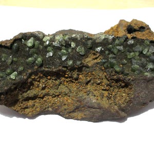 May include: A rough, irregularly shaped rock formation with a dark, textured surface. The rock features clusters of small, translucent green crystals and areas of brown and tan coloration, suggesting mineral deposits.