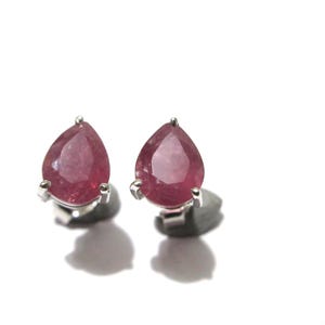 May include: A pair of stud earrings featuring faceted, pear-shaped pink gemstones set in silver-toned metal prongs. The earrings have a classic teardrop silhouette with a polished finish, showcasing the translucent quality of the pink stones.