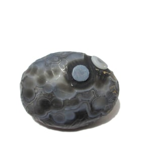May include: A polished, rounded agate stone featuring a complex pattern of translucent grey and dark charcoal orbicular inclusions. The surface displays natural concentric banding and circular formations, with two distinct, lighter-coloured circular spots.