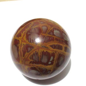 May include: A polished, spherical stone with a unique pattern. The orb features a network of golden-yellow veins against a deep reddish-brown background. The stone has a smooth, glossy surface and is set against a white backdrop.