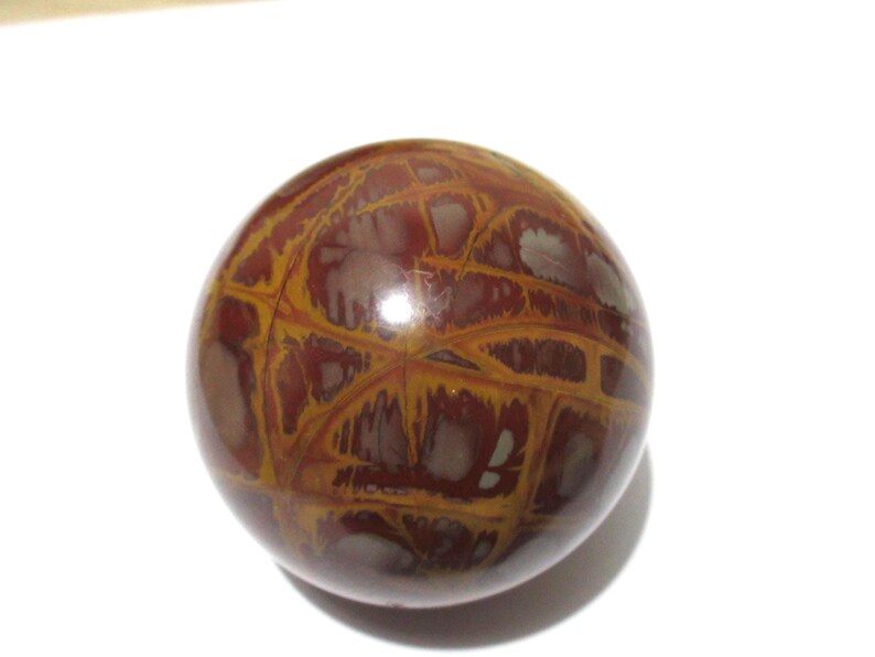 May include: A polished, spherical stone with a unique pattern. The orb features a network of golden-yellow veins against a deep reddish-brown background. The stone has a smooth, glossy surface and is set against a white backdrop.