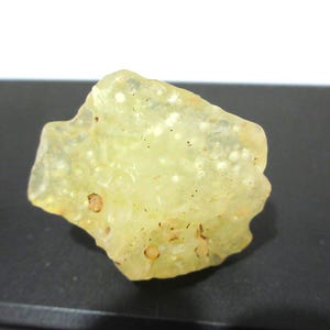 May include: A translucent, pale yellow mineral specimen with a bumpy, textured surface. The stone features small, rounded protrusions and scattered dark inclusions throughout its irregular, organic shape, displaying a semi-glossy finish.