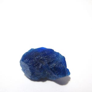 Puede incluir: Una gema en bruto, sin cortar, de color azul profundo, con una superficie rugosa y texturizada. La piedra se encuentra sobre un fondo blanco liso, resaltando su color y forma naturales. La forma irregular y el tono oscuro sugieren un zafiro o un mineral similar.