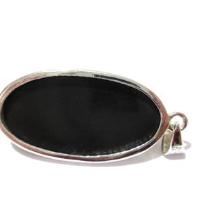 May include: Oval-shaped pendant with a polished black stone set in a silver-colored metal frame. The pendant has a loop for a chain and is against a white background. The metal is stamped with "925".