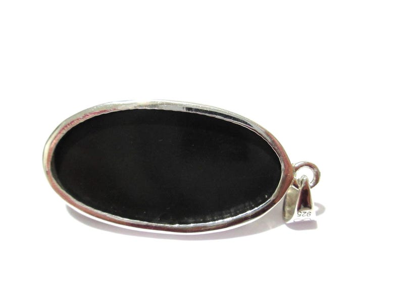 May include: Oval-shaped pendant with a polished black stone set in a silver-colored metal frame. The pendant has a loop for a chain and is against a white background. The metal is stamped with "925".