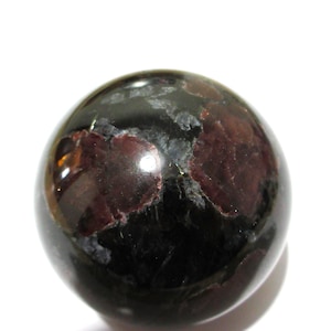 May include: A polished spherical stone featuring a deep black base colour with prominent, irregular patches of dark red and maroon mineral inclusions. The surface is smooth and reflective, showing natural variations in texture and colour throughout.
