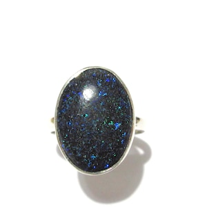 Può includere: Anello ovale con una gemma scura e macchiata, con macchie iridescenti blu e verdi, incastonato in una fascia d'argento. La gemma ha un colore di base scuro. L'anello è mostrato su sfondo bianco.