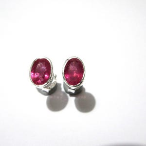 May include: A pair of oval earrings featuring bright pink gemstones set in silver. The gemstones have a polished surface. The earrings are presented against a white backdrop, showcasing their design.