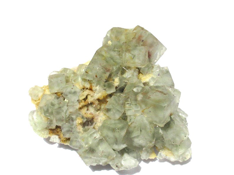 May include: A cluster of translucent, pale green fluorite crystals. The crystals have a cubic structure and are clustered together on a matrix of beige and brown minerals. The overall appearance is crystalline and natural.