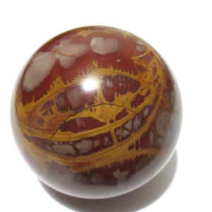 May include: A polished, spherical stone with a complex pattern of reddish-brown, yellow, and cream-colored veins and markings. The orb has a glossy finish and sits against a white background. The stone's surface is marked with a dark crack.