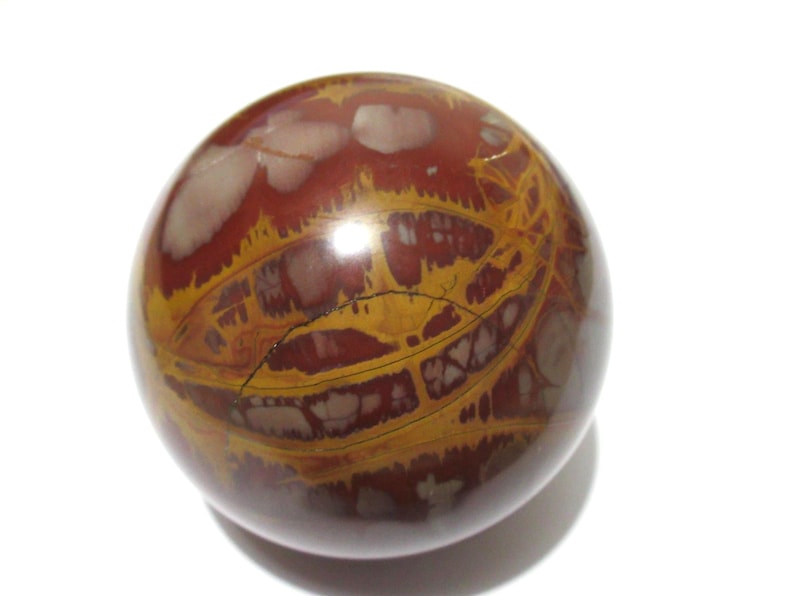 May include: A polished, spherical stone with a complex pattern of reddish-brown, yellow, and cream-colored veins and markings. The orb has a glossy finish and sits against a white background. The stone's surface is marked with a dark crack.