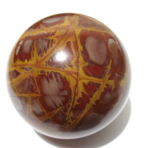 May include: A polished, spherical stone with a complex pattern. The orb features a mix of reddish-brown and yellow-gold hues, with a network of lines and shapes across its surface. The stone is set against a white background.