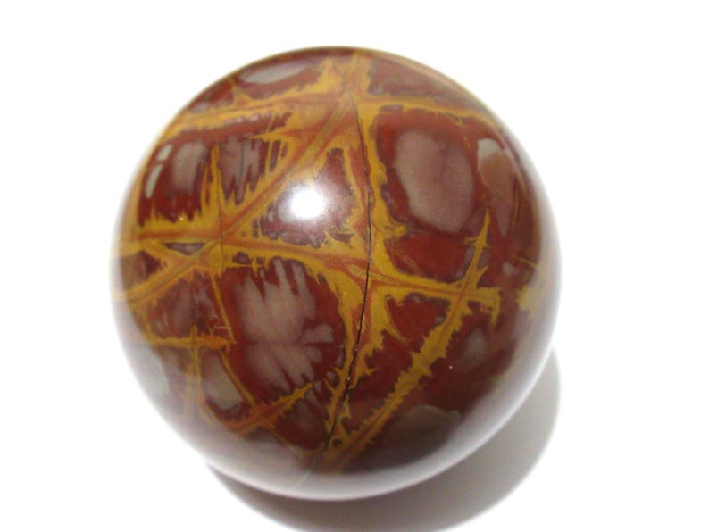 May include: A polished, spherical stone with a complex pattern. The orb features a mix of reddish-brown and yellow-gold hues, with a network of lines and shapes across its surface. The stone is set against a white background.