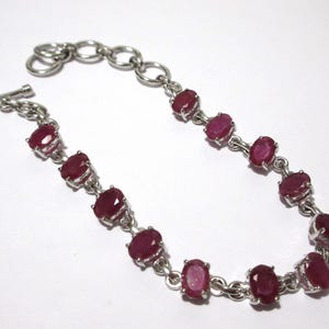 May include: A silver-toned link bracelet featuring eleven oval-shaped, deep red faceted gemstones set in individual metal frames. The bracelet includes a toggle clasp closure and a short extender chain with larger circular links at one end.
