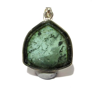 Può includere: Un ciondolo con una grande pietra preziosa verde-azzurra di forma irregolare, incastonata in una montatura in metallo color argento. La pietra presenta inclusioni scure e una superficie lucida. Il ciondolo ha un'ansa argentata.