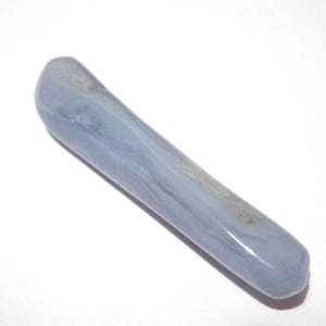 May include: A smooth, elongated blue lace agate crystal wand. The wand is a pale blue color with subtle white and gray marbling. The crystal is polished and has rounded ends. The wand is approximately 6 inches long.