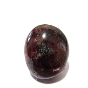 Puede incluir: Una piedra preciosa pulida, de forma ovalada, de color burdeos intenso y superficie brillante. La piedra presenta inclusiones oscuras y una ligera translucidez, sobre un fondo blanco. Es un mineral natural.