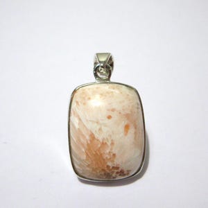 May include: A rectangular pendant featuring a polished, cream and peach-colored stone with a fibrous, fan-like internal pattern. The stone is encased in a smooth, silver-toned metal bezel setting with a small, integrated bail at the top for hanging.