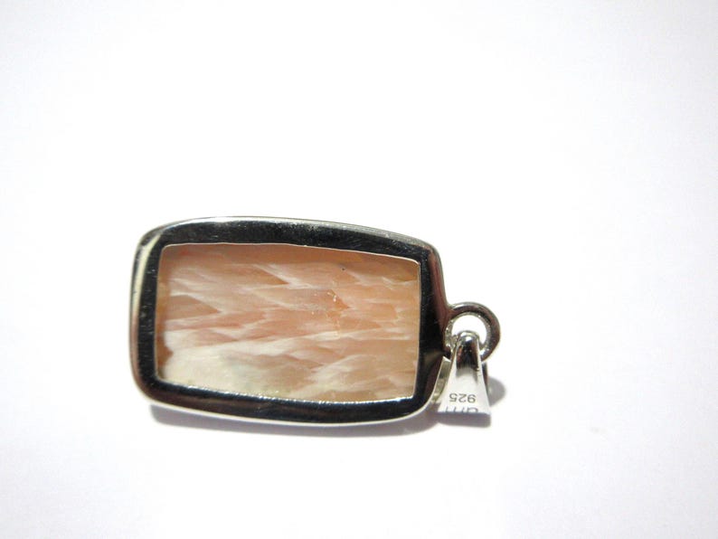 Pu&ograve; includere: Un ciondolo rettangolare con una cornice color argento. Il ciondolo presenta una pietra color pesca chiaro con un motivo fibroso. L'anello &egrave; un piccolo anello attaccato alla parte superiore della cornice. Il ciondolo &egrave; su sfondo bianco.
