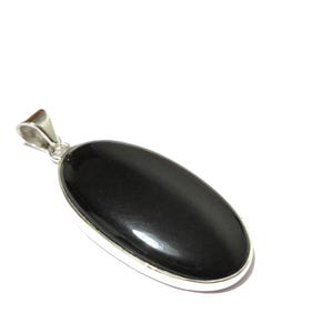 May include: A polished, oval-shaped black gemstone pendant with a silver-colored bail. The pendant is set in a silver-colored bezel. The gemstone has a smooth, glossy surface. The jewelry is displayed against a white background.