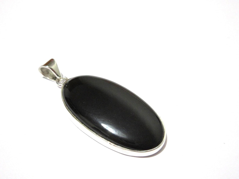 May include: A polished, oval-shaped black gemstone pendant with a silver-colored bail. The pendant is set in a silver-colored bezel. The gemstone has a smooth, glossy surface. The jewelry is displayed against a white background.