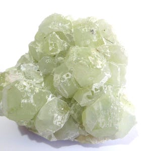 May include: A cluster of pale green crystals with a translucent appearance. The crystals have a geometric shape and are clustered together, creating a textured surface. The mineral is set against a white background.