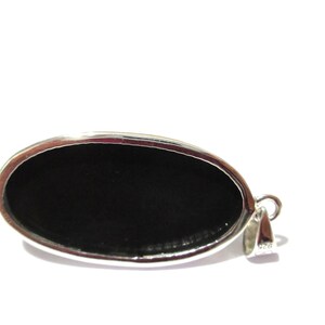 May include: Oval-shaped pendant with a polished black stone set in a silver-colored metal frame. The pendant has a loop for a chain and is set against a white background. The jewelry piece is a simple, elegant design.