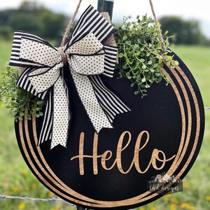 May include: A round black wooden sign with the word "Hello" in cursive script. The sign is decorated with a large bow featuring black and white striped and polka dot ribbons, and greenery. The sign is hanging by a jute rope.