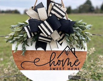 May include: Round wooden home decor sign with a white and brown design, featuring the text "home sweet home" in black script. The sign is adorned with a large black and white bow and greenery, hanging from a jute rope.