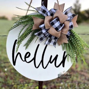 May include: A round white wooden sign with the word "hello" in black script. The sign is decorated with a large bow made of black and white checkered ribbon and burlap, along with green faux foliage. The sign is hanging from a brown rope.