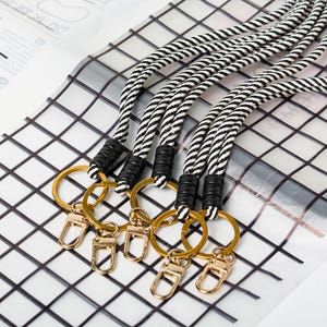 May include: Keychains with black and white striped rope lanyards. Each keychain has a gold-coloured ring and clasp, with black accents near the ring. The background features a white and grey grid pattern.