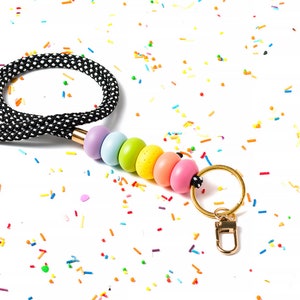 Pode incluir: Um cord&atilde;o de contas de arco-&iacute;ris com uma al&ccedil;a de bolinhas pretas e brancas e um chaveiro dourado. O cord&atilde;o est&aacute; sobre um fundo branco com confeitos coloridos.