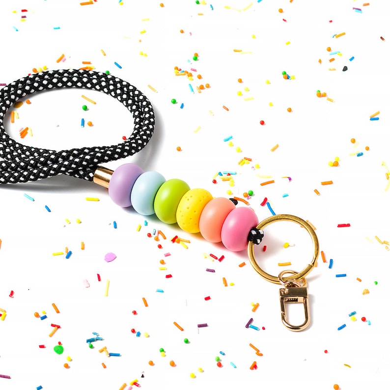 Pode incluir: Um cord&atilde;o de contas de arco-&iacute;ris com uma al&ccedil;a de bolinhas pretas e brancas e um chaveiro dourado. O cord&atilde;o est&aacute; sobre um fundo branco com confeitos coloridos.