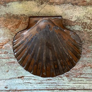 May include: A vintage, copper-toned scallop shell-shaped dish. The dish has a textured surface with radiating lines and a dark patina. The background is a distressed wooden surface with shades of brown, green, and blue.