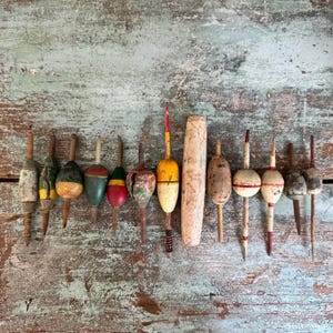May include: A collection of fourteen vintage fishing floats arranged in a horizontal row. These antique bobbers feature various shapes, including elongated, oval, and cylindrical forms, with weathered paint in shades of yellow, red, green, and white.