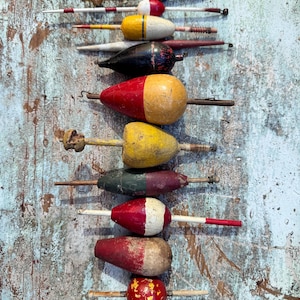 May include: A collection of twelve vintage fishing floats arranged vertically. These wooden and plastic bobbers feature various shapes, including spheres, teardrops, and cylinders, painted in red, yellow, white, black, green, and blue striped patterns.