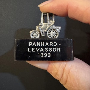 May include: A silver miniature car model with spoked wheels and a carriage-like body, mounted on a black rectangular base. The base features the text "PANHARD - LEVASSOR 1893" in white lettering. The model is held against a dark background.