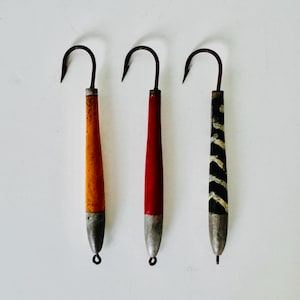 May include: Three fishing lures arranged vertically, each featuring a curved metal hook at the top, a cylindrical body with distinct finishes—one orange, one red, and one black with white stripes—and a weighted metal tip with a small loop at the base.