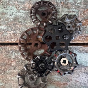 May include: A collection of vintage metal valve handles in various sizes and colors, including rusty brown, black, and silver. The handles have a circular design with spokes and a central square opening, suggesting an industrial or steampunk theme.