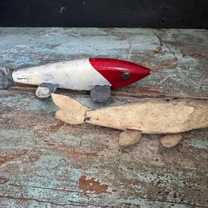 May include: Two wooden fish sculptures. One is painted white with a red head and a gray tail fin. The other is a natural wood color. Both have small, gray wheels attached to the bottom.