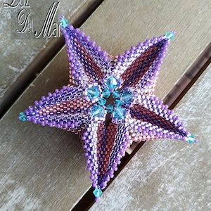 May include: A purple and pink beaded star with a turquoise centre. The star is made with seed beads and has a 3D effect. The star is on a wooden surface.