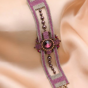 May include: A beaded bracelet with a pink and silver design. The bracelet features a large, faceted, purple gemstone in the centre. The bracelet is on a pink fabric background.