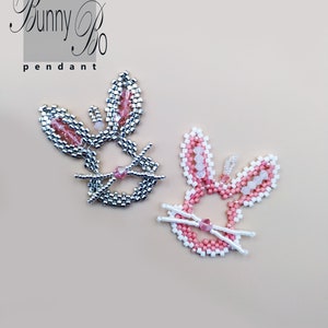 Può includere: Due ciondoli di coniglio perlati, uno con perline argentate e rosa, l'altro con perline bianche e rosa. I ciondoli sono su uno sfondo marrone chiaro. Il testo "Bunny Bo pendant" è in alto nell'immagine.