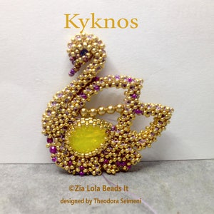 Könnte beinhalten: Ein goldener und lila Perlenswan mit einem gelben Mittelstück. Der Schwan ist ein Schmuckstück aus Perlen mit dem Titel "Kyknos" darüber und dem Text "© Zia Lola Beads It designed by Theodora Seimeni" darunter.