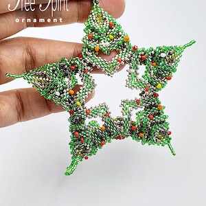 Könnte beinhalten: Ein sternförmiger Anhänger aus grünen, silbernen und roten Perlen mit dem Text "Tree Spirit ornament" darüber. Der Anhänger wird in einer Hand gehalten.